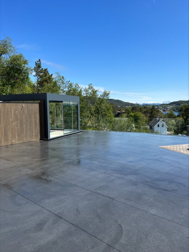 Flott takterrasse med ny støpt betongflate og glassrekkverk, omgitt av grøntområder.